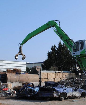 Grue de manutention SENEB verte saisissant des matériaux avec sa pince, sur un site de ferraille avec des carcasses de voitures écrasées.