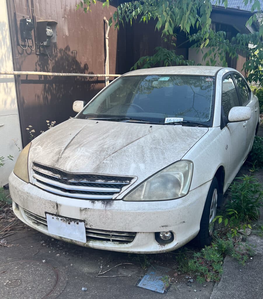 Voiture Toyota Allion sale et abandonnée dehors Toyota Allion blanche très sale, stationnée à l'extérieur près d'un mur brun avec des compteurs électriques et envahie par la végétation.